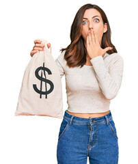 Sticker - Young brunette woman holding dollars bag covering mouth with hand, shocked and afraid for mistake. surprised expression