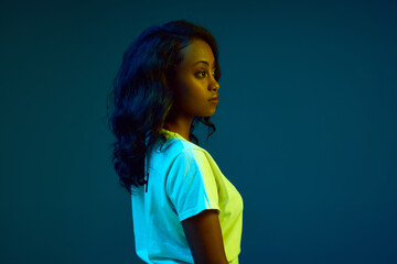 Profile portrait of attractive young, african woman in white t-shirt posing against cyan, blue studio background in neon light. Concept of human emotions, youth, fashion, lifestyle, ad