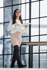 Beautiful confident young busines woman with black hair in a modern office standing near the table with a folder in her hand