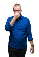 Poster - Young caucasian hipster man wearing glasses over isolated background smelling something stinky and disgusting, intolerable smell, holding breath with fingers on nose. Bad smells concept.