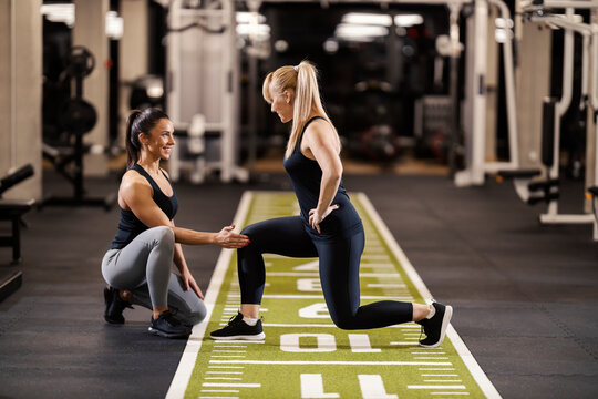 A happy female personal instructor is helping sportswoman with lunges in a gym.