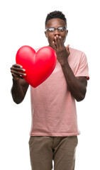 Canvas Print - Young african american man holding red heart cover mouth with hand shocked with shame for mistake, expression of fear, scared in silence, secret concept