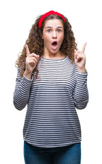 Poster - Beautiful brunette curly hair young girl wearing stripes sweater over isolated background amazed and surprised looking up and pointing with fingers and raised arms.