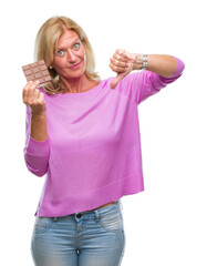 Canvas Print - Middle age blonde woman eating chocolate bar over isolated background with angry face, negative sign showing dislike with thumbs down, rejection concept