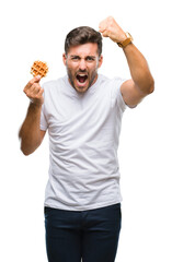 Wall Mural - Young handsome man eating a sweet waffle over isolated background annoyed and frustrated shouting with anger, crazy and yelling with raised hand, anger concept