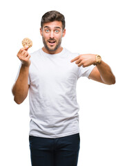 Canvas Print - Young handsome man eating chocolate chips cookie over isolated background with surprise face pointing finger to himself