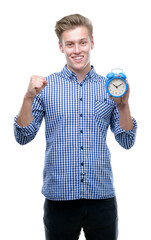 Wall Mural - Young handsome blond man holding alarm clock screaming proud and celebrating victory and success very excited, cheering emotion