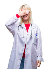 Poster - Young beautiful blonde doctor woman wearing medical uniform over isolated background smiling and laughing with hand on face covering eyes for surprise. Blind concept.