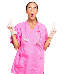 Wall Mural - Young hispanic woman wearing doctor uniform and stethoscope amazed and surprised looking up and pointing with fingers and raised arms.