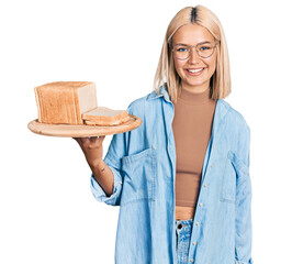 Sticker - Beautiful young blonde woman holding bread looking positive and happy standing and smiling with a confident smile showing teeth