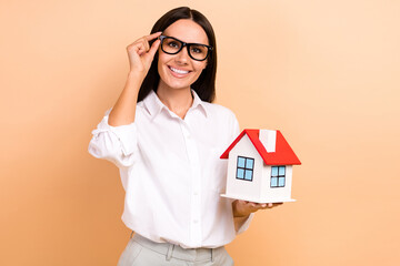 Wall Mural - Portrait of pretty positive realtor lady arm touch eyeglasses hold little house isolated on beige color background