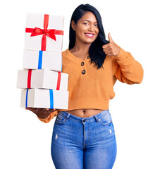 Poster - Hispanic woman with long hair holding gifts smiling happy and positive, thumb up doing excellent and approval sign