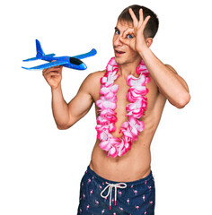 Wall Mural - Young blond man wearing swimsuit and hawaiian lei holding airplane toy smiling happy doing ok sign with hand on eye looking through fingers