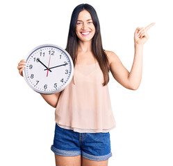 Canvas Print - Young beautiful caucasian woman holding big clock smiling happy pointing with hand and finger to the side