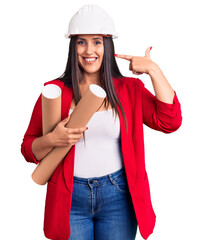 Poster - Young beautiful brunette woman wearing hardhat holding architect blueprint pointing finger to one self smiling happy and proud