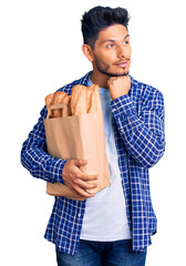 Sticker - Handsome latin american young man holding paper bag with bread with hand on chin thinking about question, pensive expression. smiling and thoughtful face. doubt concept.