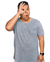 Poster - Young latin man wearing casual clothes doing ok gesture shocked with surprised face, eye looking through fingers. unbelieving expression.