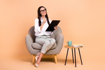 Full length photo of funny dreamy lady wear white shirt sitting chair writing documents looking empty space isolated beige color background