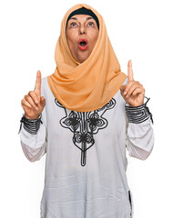 Wall Mural - Middle age hispanic woman wearing traditional islamic hijab scarf amazed and surprised looking up and pointing with fingers and raised arms.