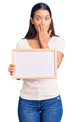 Canvas Print - Young beautiful latin girl holding empty white chalkboard covering mouth with hand, shocked and afraid for mistake. surprised expression