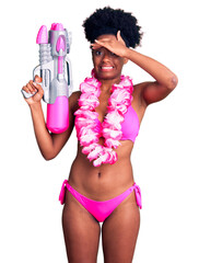 Poster - Young african american woman wearing bikini and hawaiian lei holding water gun stressed and frustrated with hand on head, surprised and angry face