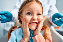 Humorous Portrait Of A Cute Little Girl With Pigtails Sitting In A Dental Chair, Looking At The Camera And Grimacing. Behind, A Doctor In Gloves Holds Examination Tools.