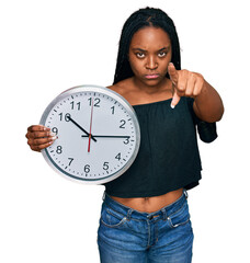 Sticker - Young african american woman holding big clock pointing with finger to the camera and to you, confident gesture looking serious