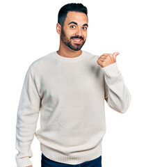 Poster - Young hispanic man with beard wearing casual white sweater smiling with happy face looking and pointing to the side with thumb up.