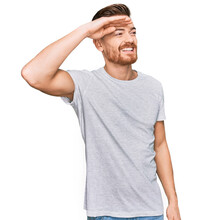 Young Redhead Man Wearing Casual Grey T Shirt Very Happy And Smiling Looking Far Away With Hand Over Head. Searching Concept.