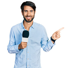 Hispanic man with blue eyes holding reporter microphone smiling happy pointing with hand and finger to the side