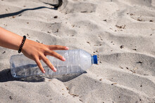 A Hand Picking Up An Empty Plastic Bottle From The Sand On The Beach, Ecology Concept