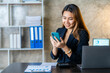 © ArLawKa - Asian businesswoman holding smartphone in talking with business clients with charts or documents in workplace using calculator, business laptop, financial accountant calculating money, loan bank,