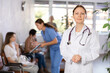© JackF - Smiling polite female doctor in white coat posing indoors against blurred background of busy clinic lobby with healthcare workers and patients..