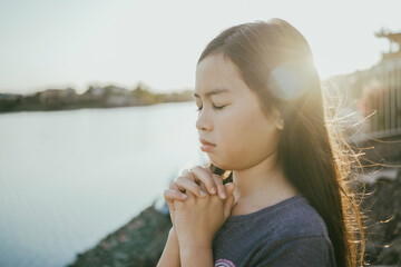 Wall Mural - Preteen mixed girl praying with eyes closed by lake, kid worshiping online at home, peace and hope concept