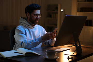 Canvas Print - Home workplace. Happy man using smartphone while working with computer at wooden desk in room at night