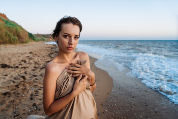 Wall Mural - a young woman stands on the seashore wrapped in a cloth at sunset