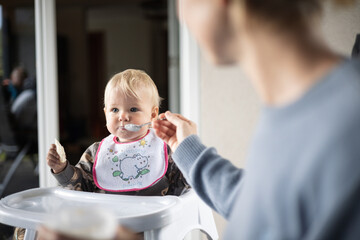 Wall Mural - Mother spoon feeding her baby boy child in baby high chair with fruit puree at dinning table at home. Baby solid food introduction concept.