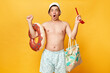 Amazed man wearing shorts swimsuit and panama holding donut rubber ring isolated on yellow background looking at camera with big eyes keeps mouth open raised arms.
