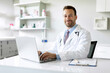 Portrait of smiling european doctor wearing white uniform sitting at desk with laptop smiling at camera, man GP or physician working in private clinic