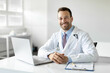 © Home-stock - Portrait of cheerful man doctor in workwear working in modern clinic, sitting at workdesk with laptop and medical chart smiling at camera