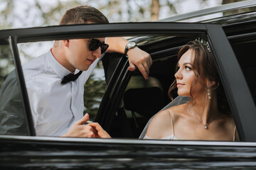 Wedding portrait, photo of a stylish groom in a white shirt and bow tie and a brunette bride with a bouquet of flowers near a black car.