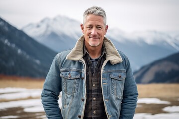 Wall Mural - Studio portrait photography of a tender mature man wearing a denim jacket against a serene snow-capped mountain background. With generative AI technology