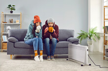Full Length Photo Of A Young Frozen Family Couple Man And Woman Sitting On Sofa In The Living Room In Winter Outerwear And In Hats At Home Warming Their Hands. Heating Problems Concept.