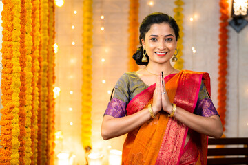 Happy indian girl with traditional ethnic dress showing namaste gesture by looking at camera during festival celebration at home - concept of welcome, greetings and Indian culture.