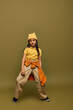 Full length of confident preadolescent girl in urban outfit and yellow hat posing and looking at camera while standing on khaki background, stylish girl in modern outfit concept