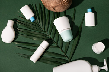 Still life. Flat lay composition with set of white bottles with body lotion, moisturizer, facial anti-aging serum laid out on a palm leaf, isolated over dark green background. Brand packaging mockup