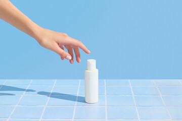 Poster - Womans hand holding white tube on blue tile background. Beauty treatment. Skin care routine