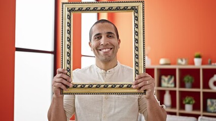 Wall Mural - Young hispanic man smiling confident holding empty frame at home