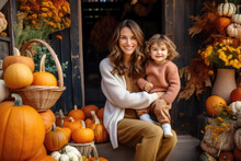 Happy Young Woman Sitting On The Stairs With Her Little Daughter, On A Porch Beautifully Decorated With Autumn Pumpkins 