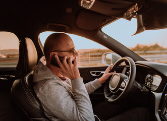 A man with a sunglasses driving a car and talking on smartphone at sunset. The concept of car travel